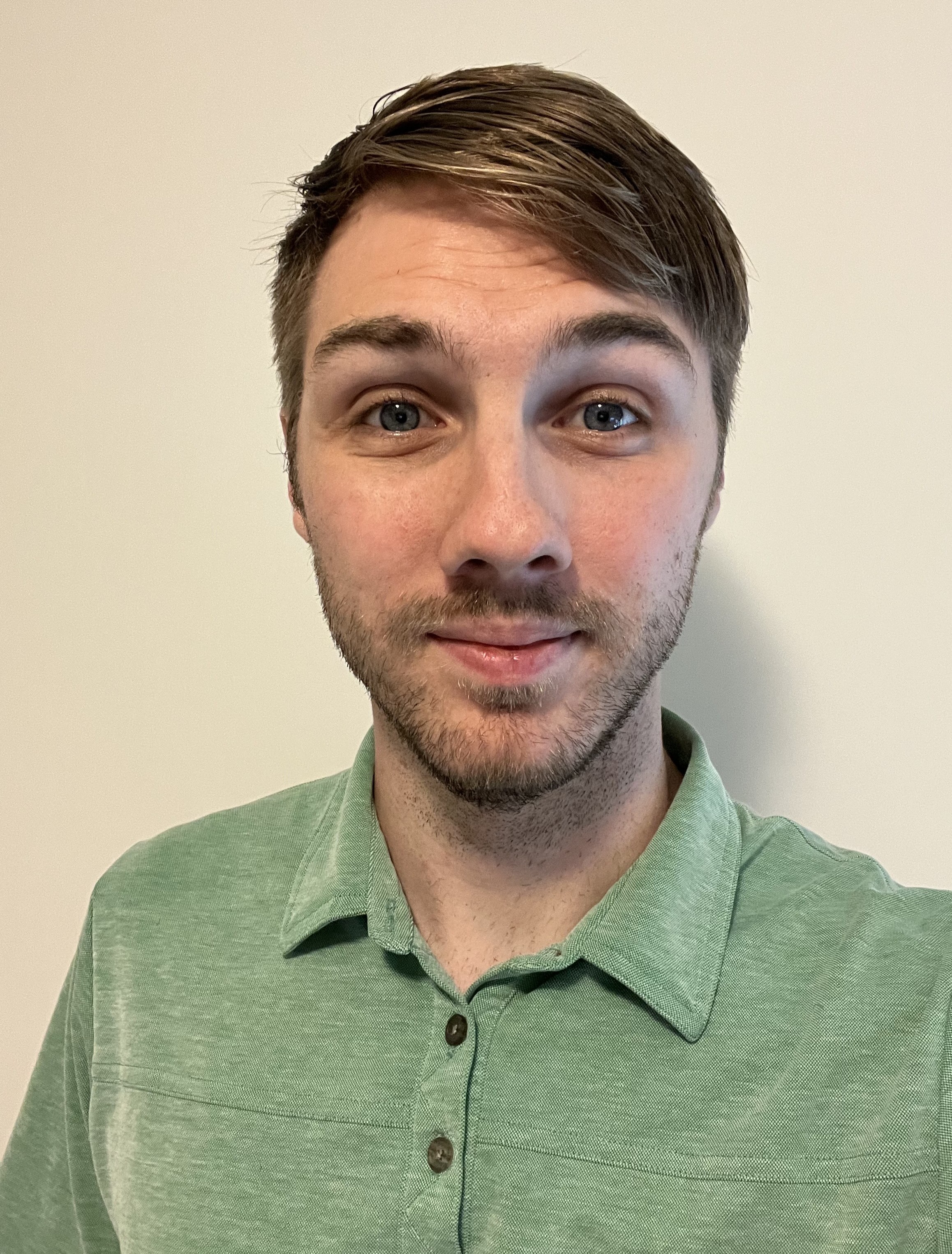Young man with short brown hair and light beard wearing a green collared shirt, smiling slightly in front of a plain beige wall.